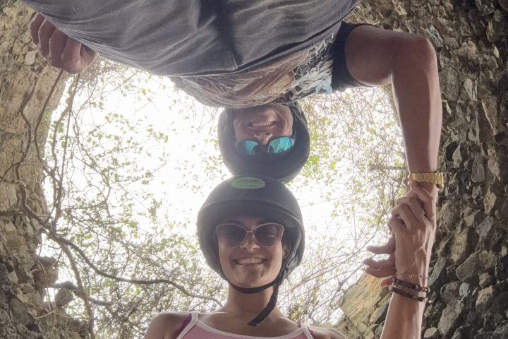Two people wearing helmets stand in a circular stone structure, holding hands and looking down.