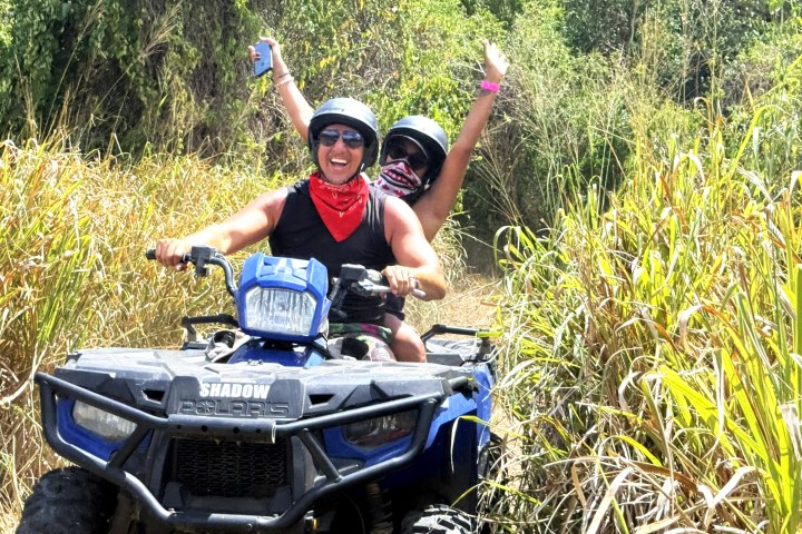 Two people on an ATV, smiling and raising arms, surrounded by tall grass.