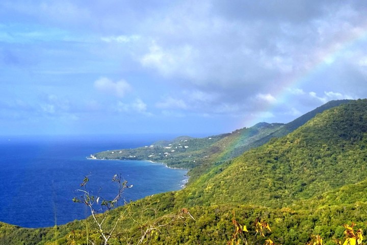 Coastal landscape with green hills, blue sea, and a faint rainbow in the cloudy sky.